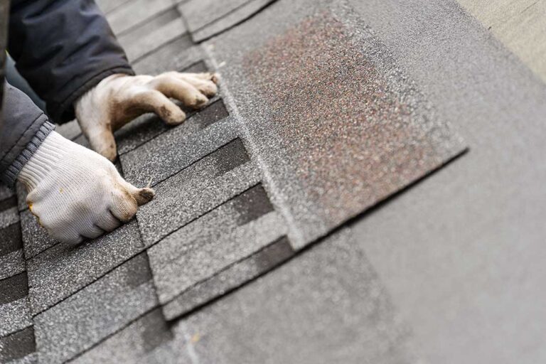 Person inspecting roof gutters from a ladder beside a home with leaves and autumn debris nearby.